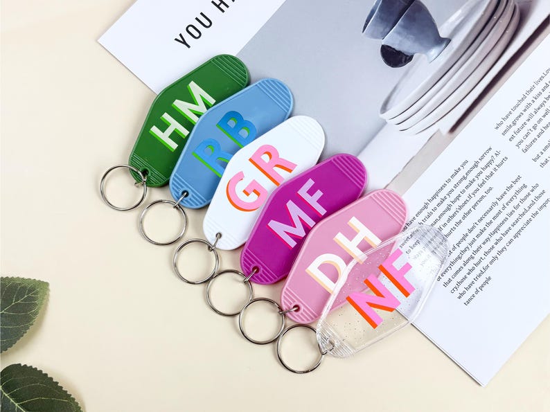 K&ouml;nnte beinhalten: Eine Sammlung farbenfroher Schl&uuml;sselanh&auml;nger in Gr&uuml;n, Blau, Wei&szlig;, Lila, Rosa und Transparent. Jeder Schl&uuml;sselanh&auml;nger hat ein einzigartiges Design mit zwei Buchstaben in kr&auml;ftigen, kontrastierenden Farben. Die Schl&uuml;sselanh&auml;nger sind an silbernen Schl&uuml;sselringen befestigt.