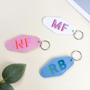 K&ouml;nnte beinhalten: Drei Schl&uuml;sselanh&auml;nger im Retro-Stil in Rosa, Wei&szlig; und Blau. Jeder Schl&uuml;sselanh&auml;nger hat unterschiedliche Initialen in einer Kontrastfarbe: "MF", "NF" und "RB". Die Schl&uuml;sselanh&auml;nger sind an silbernen Schl&uuml;sselringen befestigt.