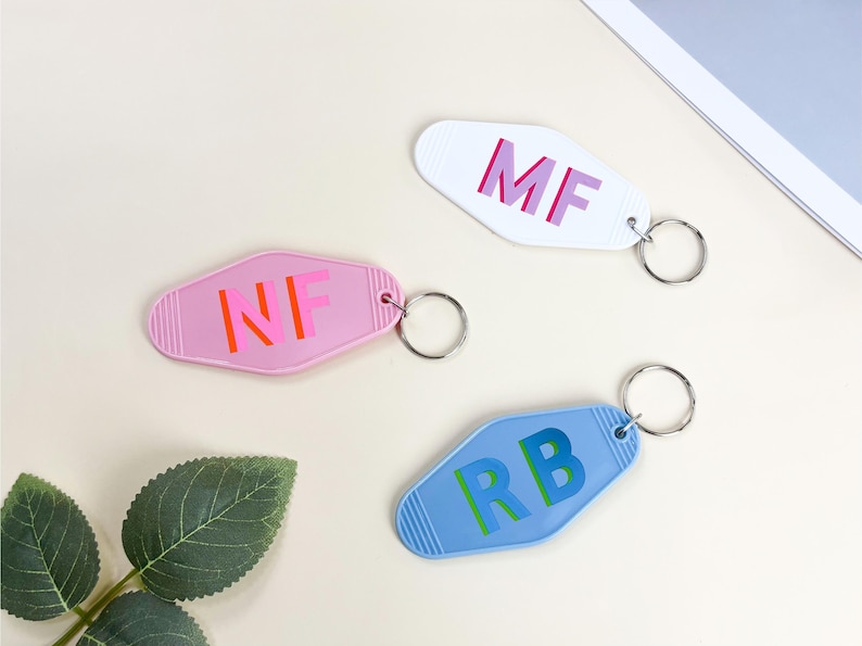 K&ouml;nnte beinhalten: Drei Schl&uuml;sselanh&auml;nger im Retro-Stil in Rosa, Wei&szlig; und Blau. Jeder Schl&uuml;sselanh&auml;nger hat unterschiedliche Initialen in einer Kontrastfarbe: "MF", "NF" und "RB". Die Schl&uuml;sselanh&auml;nger sind an silbernen Schl&uuml;sselringen befestigt.