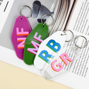K&ouml;nnte beinhalten: Vier farbige Schl&uuml;sselanh&auml;nger mit silbernen Ringen. Die Schl&uuml;sselanh&auml;nger sind in Lila, Gr&uuml;n, Wei&szlig; und Transparent gehalten und tragen die Buchstaben NF, MF, RB und GR in verschiedenen Farben. Auf einer wei&szlig;en Oberfl&auml;che.