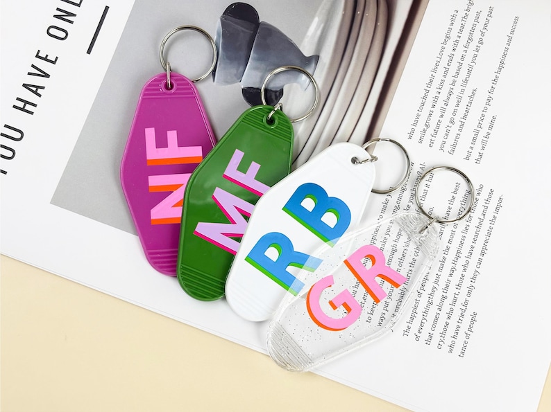 K&ouml;nnte beinhalten: Vier farbige Schl&uuml;sselanh&auml;nger mit silbernen Ringen. Die Schl&uuml;sselanh&auml;nger sind in Lila, Gr&uuml;n, Wei&szlig; und Transparent gehalten und tragen die Buchstaben NF, MF, RB und GR in verschiedenen Farben. Auf einer wei&szlig;en Oberfl&auml;che.