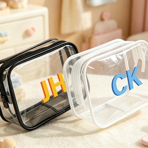 Könnte beinhalten: Zwei transparente Kosmetiktaschen mit schwarzen und weißen Rändern. Eine Tasche hat die Initialen "JH" in Orange, die andere "CK" in Blau. Beide Taschen haben einen Reißverschluss und sind rechteckig.