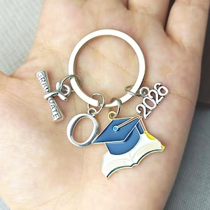 May include: Silver graduation keychain with a mortarboard, open book, diploma, and the year 2026. The mortarboard is blue and gold. The diploma reads "GRADUATE". The keychain is held in a hand.