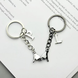 May include: Two keychains connected by a chain, each with a silver ring. One keychain has the letter "P" and the other has the letter "L". Both keychains have a charm of two hands holding each other.