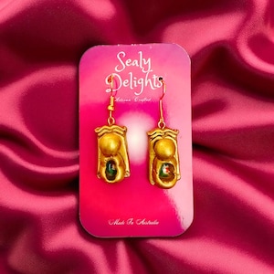 Könnte beinhalten: Goldfarbene Ohrringe mit rechteckigem Design und einem kleinen, farbigen Edelstein. Die Ohrringe werden auf einer rosa Karte mit dem Text "Sealy Delights" und "Made In Australia" präsentiert.