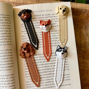 May include: Five dog-themed bookmarks with detailed sculpted dog heads. The bookmarks feature a Rottweiler, Cavalier King Charles Spaniel, Shiba Inu, Golden Retriever, and Husky. Each bookmark is a different color, and they are resting on the pages of a book.