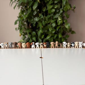 Può includere: Una collezione di piccole statuette di cani di varie razze e colori, disposte in fila su una superficie bianca. Le statuette sono dettagliate e realistiche, con diverse pose e segni. Una pianta verde sullo sfondo.