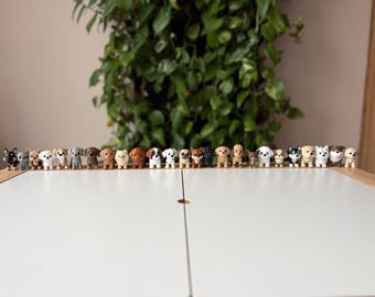 May include: A collection of small dog figurines in various breeds and colors, arranged in a row on a white surface. The figurines are detailed and realistic, with different poses and markings. A green plant is in the background.
