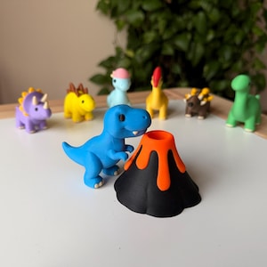 May include: A collection of colorful dinosaur figurines, including a blue Tyrannosaurus Rex, a purple Triceratops, and a yellow dinosaur. A black volcano with orange lava is in the foreground. The toys are on a white surface.