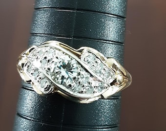May include: A gold ring with a cluster of diamonds. The ring features a central, larger diamond surrounded by smaller stones, set in a decorative, flowing design. The band is a polished gold color.