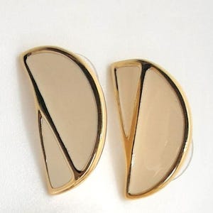 May include: A pair of gold-toned stud earrings. Each earring features a half-circle design with a cream-colored enamel filling and a black accent line. The earrings have a post backing.