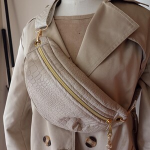 Peut inclure: Une banane en similicuir beige avec une fermeture éclair dorée et un charme de chaîne dorée. La banane est portée sur un trench beige.