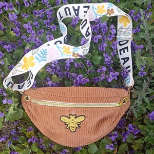 Peut inclure: Une banane en velours côtelé marron avec une fermeture éclair dorée et une abeille brodée sur le devant. La banane a une sangle blanche avec des motifs floraux et des feuilles et les mots "IDEAL" et "BEAUT" imprimés dessus.