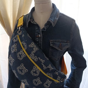 以下が含まれることがあります： 黄色のジッパーと白い虎のプリントが施された青いデニムのウエストバッグ。デニムジャケットに着用されています。