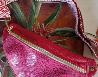 May include: A pink snakeskin fanny pack with a gold zipper and a red and white patterned strap.