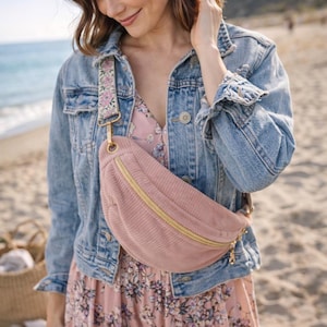 Peut inclure: Un sac banane en velours côtelé rose avec une fermeture éclair et des accessoires dorés. Le sac a une sangle florale et est porté sur une robe fleurie et une veste en jean. Le fond est une scène de plage.