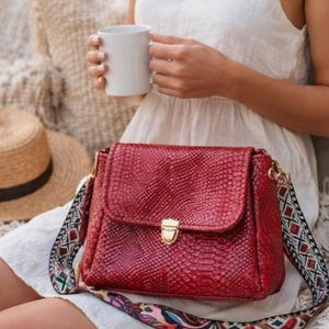 May include: A red handbag with a textured, snake-like pattern. The bag has a gold-colored clasp and a colorful patterned strap. A straw hat and a white mug are also visible.