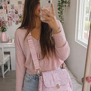 May include: A light pink crossbody bag with a textured pattern and gold-colored hardware. The bag has a matching pink strap with a repeating logo design. The person is wearing a light pink sweater and blue jeans.