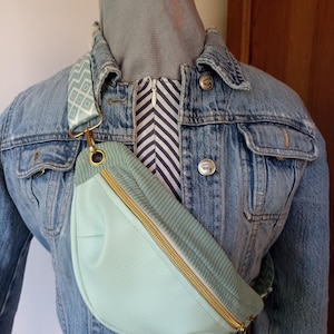 Könnte beinhalten: Eine hellgrüne Bauchtasche mit einem goldenen Reißverschluss und einem weißen und grünen gemusterten Gurt. Die Bauchtasche wird über einer Jeansjacke getragen.