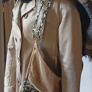 Puede incluir: Un bolso bandolera de pana marrón y estampado de leopardo con una correa de cadena dorada. El bolso lo lleva un maniquí que lleva una gabardina beige y una bufanda negra y marrón.