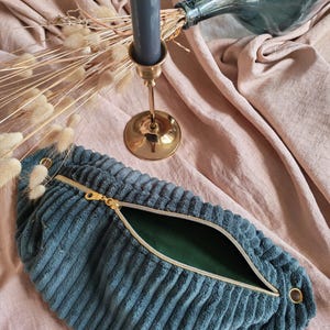 Peut inclure: Sac banane en velours côtelé bleu canard avec une fermeture éclair dorée et un intérieur vert. Le sac est ouvert, révélant l'intérieur. Le fond comprend un bougeoir doré, une bougie grise et une bouteille en verre avec des fleurs séchées.