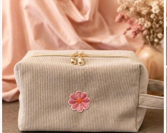 May include: A rectangular beige corduroy toiletry bag featuring a gold-toned double zipper closure and a small embroidered pink flower with an orange center on the front. The bag includes a matching side handle made of the same corduroy fabric.