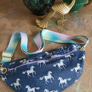 Peut inclure: Une banane en jean bleu avec un imprimé de cheval blanc et une sangle réglable bleu clair et rose. La banane a une fermeture éclair.