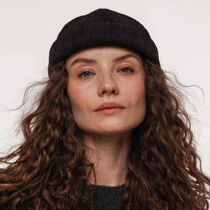 May include: A black ribbed knit beanie. The beanie is worn on the head. The person is wearing a black coat and a dark green jumper. The person has long, curly brown hair and is looking at the camera.