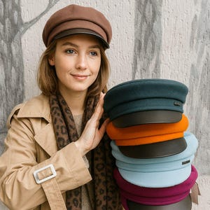 May include: A selection of newsboy caps in various colours: teal, orange, light blue, and burgundy, each with a dark brown brim. A woman holds the caps, wearing a brown cap, a tan trench coat, and a leopard print scarf.