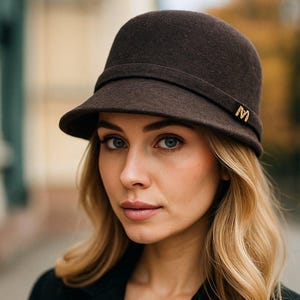 Può includere: Un cappello cloche marrone scuro con una piccola tesa e un'emblema "M" dorato. Il cappello è indossato con un cappotto doppiopetto nero e un maglione beige. La donna ha i capelli biondi ondulati e guarda la fotocamera.