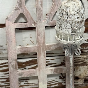 May include: A distressed, pink-painted wooden window frame with a decorative, white lace-covered birdcage on a metal stand. The window frame has a weathered appearance, and the birdcage is suspended from a curved metal handle.