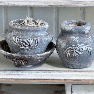 May include: Two small, weathered gray pots with a textured, stone-like finish. One pot sits inside a matching saucer, while the other stands alone. Both feature raised, white floral relief detailing on their rounded bodies and wide rims.