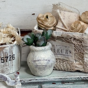 May include: A small, white, distressed ceramic vase with green and blue faux flowers. The vase sits on a light blue surface next to a white wooden box with the number 5726 printed on it. A basket with fabric roses is in the background.