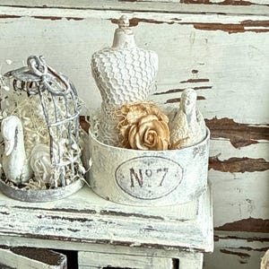 May include: A white distressed metal container labeled No 7 containing a miniature dress form and a fabric rose. Beside it sits a small white wire birdcage with a swan figurine inside, both featuring a weathered, aged finish.