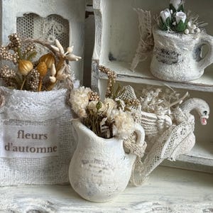 May include: A collection of miniature white decorative items including a fabric pouch labeled 'fleurs d'automne', a small pitcher with dried flowers, a swan figurine adorned with lace, and a mug containing white roses, all set on a white shelf.