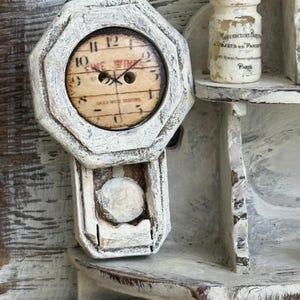 Puede incluir: Un reloj de madera blanca envejecida con marco octogonal. La esfera del reloj es de color marrón claro con números y manecillas negras. Un pequeño jarrón blanco con flores secas está sobre una estantería. La palabra "forever" es visible.