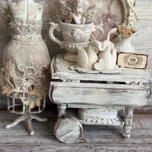May include: A collection of vintage-style decorative items in shades of white and cream. Includes a miniature dress form, a small cart, a teacup with an angel, swan figurines, a picture frame, and a small vase. The items have a distressed, antique appearance.