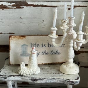 May include: A miniature white lighthouse with a pink top sits on a small, distressed white table. Beside it is a white candelabra with lit candles. A small sign reads "life is better by the lake."