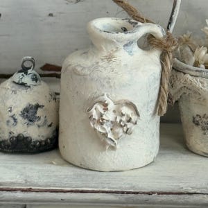 May include: A small, off-white ceramic jug with a heart-shaped embellishment and a jute rope handle. The jug is part of a collection of decorative items, including a bell-shaped ornament and a small bucket with floral accents.
