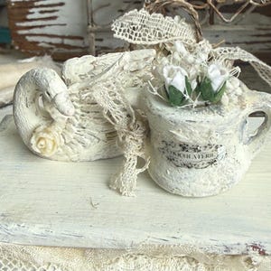 May include: A decorative off-white swan figurine attached to a small watering can, featuring a distressed paint finish, a small cream-colored rose on the swan, and two white fabric rosebuds with green leaves inside the watering can opening.