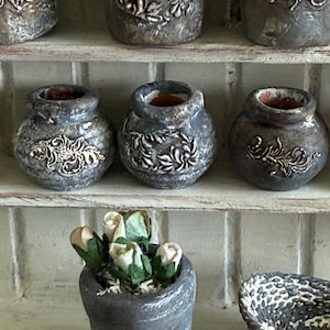 May include: A collection of small, dark gray, textured ceramic pots and vases featuring raised white floral relief patterns. Some pots are empty, while one contains a cluster of small, pale, cream-colored artificial rose buds.