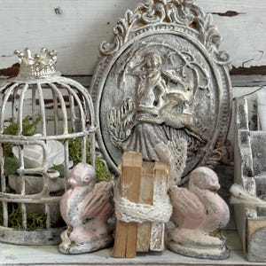 May include: A decorative display featuring a white birdcage with a crown, a detailed oval relief, and two pink and gray bird figurines. A bundle of wooden sticks is wrapped with white twine.