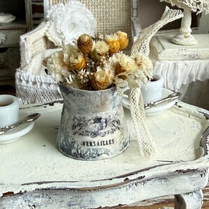 May include: A miniature metal bucket with a distressed white finish and the word VERSAILLES printed on the front. The bucket is filled with a bouquet of dried yellow and cream-colored flowers and features a decorative piece of white lace ribbon.