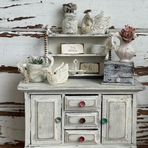 May include: A miniature white wooden cabinet with three central drawers featuring floral knobs and two side doors. The top surface holds a small hutch displaying a swan figurine, a pitcher with pink flowers, a sign reading dream, and a sign reading No. 6.