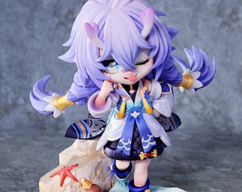 May include: A small, purple-haired figurine of a character from the video game Star Rail. The figurine is wearing a white and blue outfit with a gold belt and is standing on a sandy base with seashells and starfish.