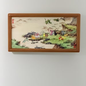 May include: A framed artwork with a brown wooden frame, depicting a landscape with cartoon characters. The scene shows a green landscape with mountains, water, and various colorful cartoon characters. The style is reminiscent of traditional Asian art.