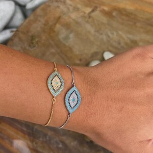 Könnte beinhalten: Zwei verstellbare Armbänder mit einem blauen bösen Augen-Anhänger. Die Anhänger sind mit klaren Strasssteinen besetzt. Ein Armband hat eine goldene Kette und das andere eine silberne Kette.