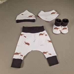 Könnte beinhalten: Ein Baby-Outfit-Set mit einer weißen Mütze, einem Lätzchen und einer Hose mit einem braunen Schildkröten-Druck. Die Hose hat dunkelgraue Bündchen und einen dunkelgrauen Bund. Das Lätzchen hat einen dunkelorangen Saum. Das Set enthält auch ein Paar braune und weiße Stiefelchen.
