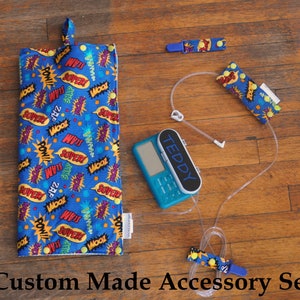 May include: A custom-made accessory set for a medical pump, featuring a blue fabric pouch with a comic book print, two blue and yellow fabric straps with snaps, and a clear tubing. The pump is blue and has the word "TEDDY" printed on it.