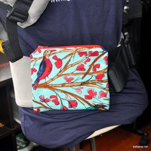 May include: A small, square pouch with a red zipper and a colorful bird and floral pattern. The background is a light blue with brown branches and red flowers. The pouch is attached to a baby carrier.